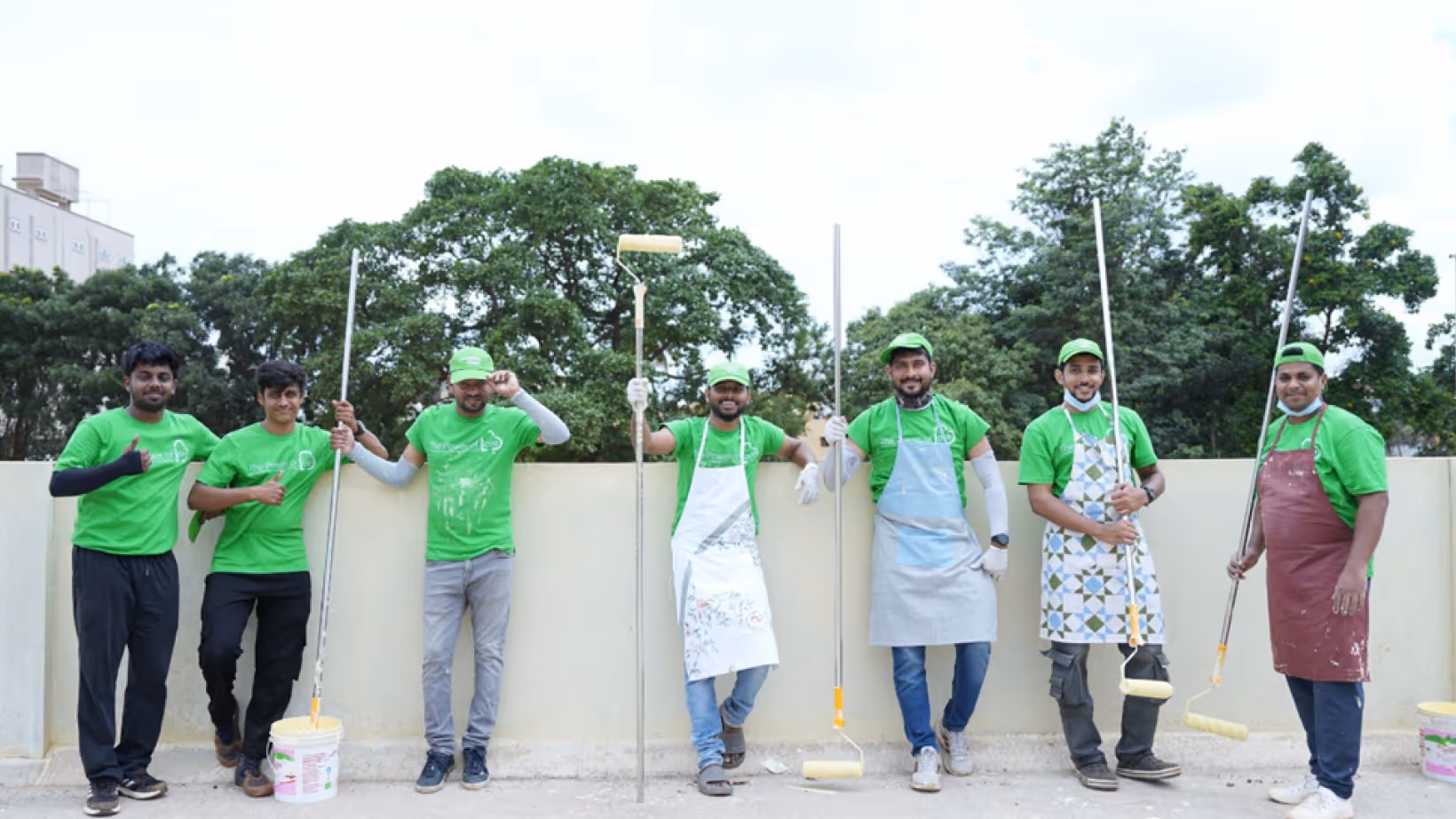 Seven men in green shirts and caps holding paint rollers and buckets, posing with thumbs up outdoors.
