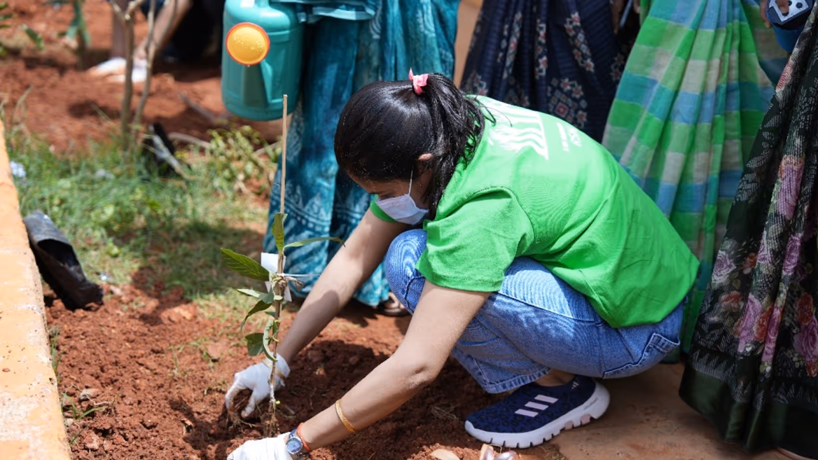 Person wearing a green shirt and gloves planting a small tree seedling in red soil outdoors.