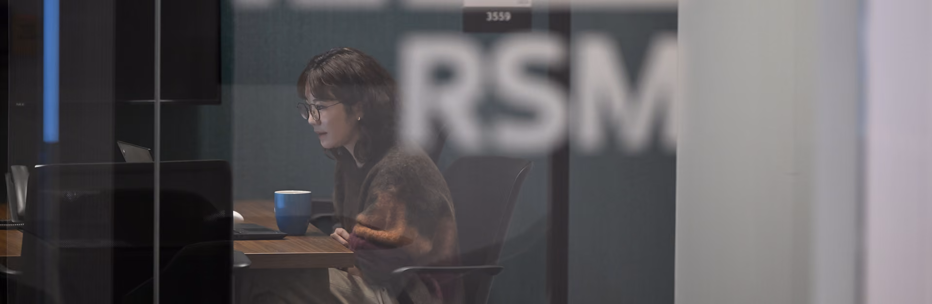 Woman wearing glasses working on a laptop at a desk with a blue coffee mug nearby.