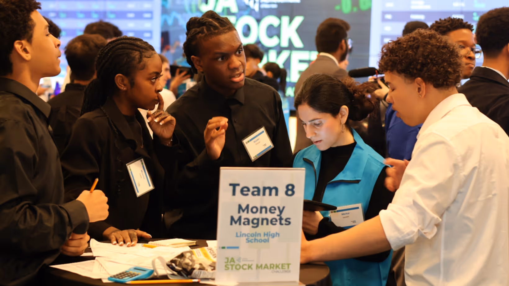 High school students in discussion at a stock market challenge event with papers and calculator on table.