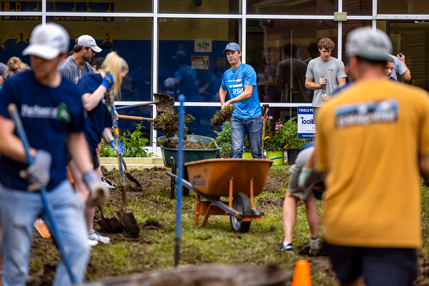 Volunteers using shovels and a wheelbarrow to dig and work in a garden outside a building.