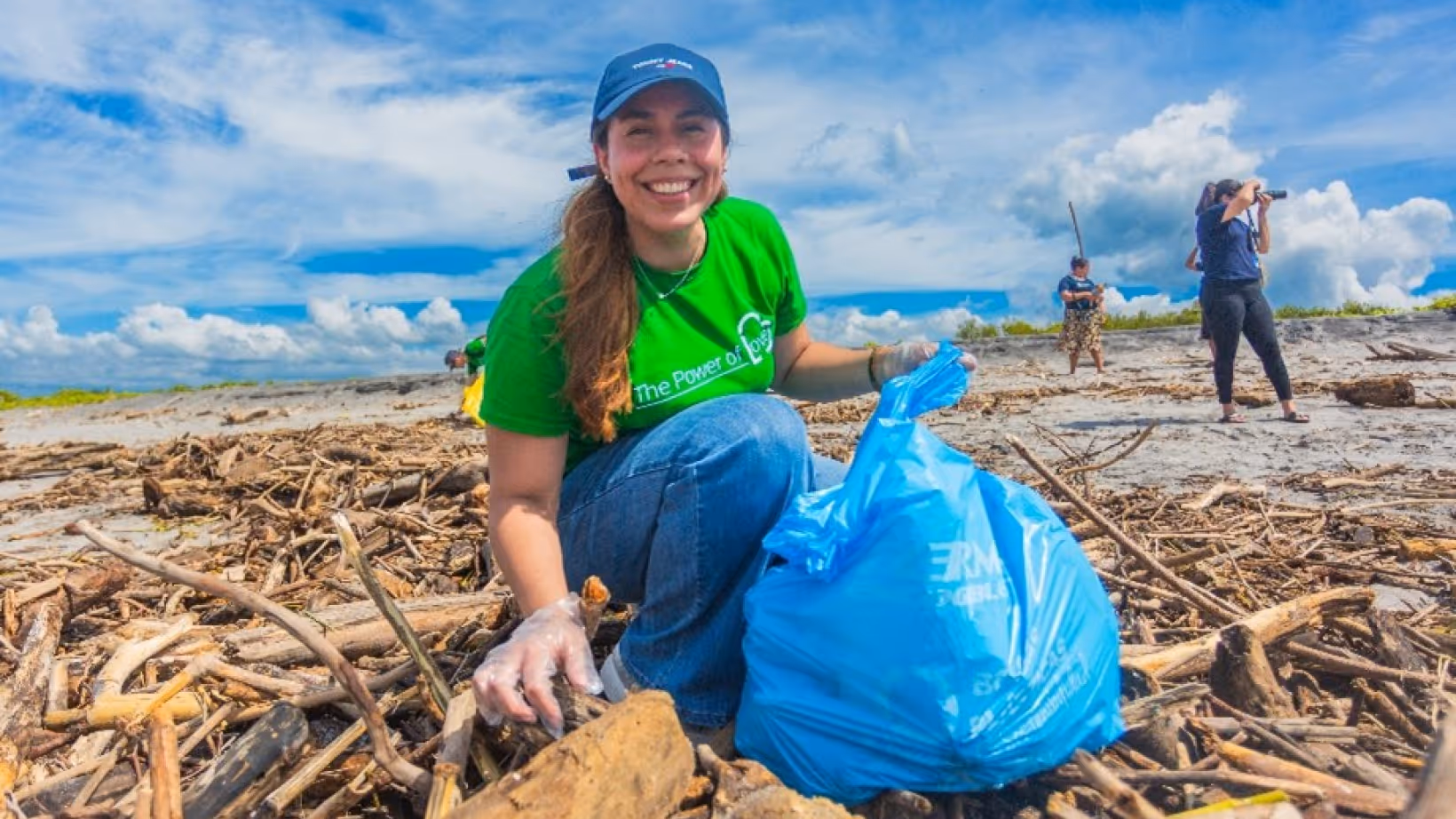 Smiling woman in green shirt and blue hat picking up trash on a beach with a blue garbage bag.