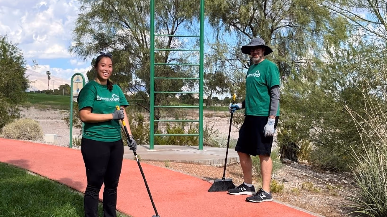 Two volunteers in green shirts holding brooms standing on a red walking path outdoors.