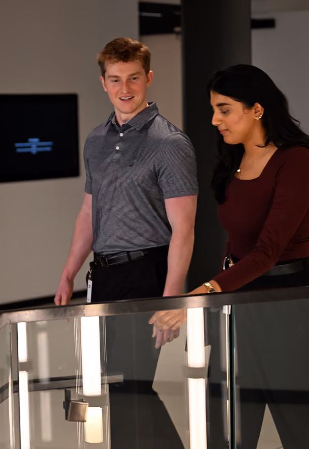 Young man and woman walking together indoors near a glass railing, engaged in conversation.