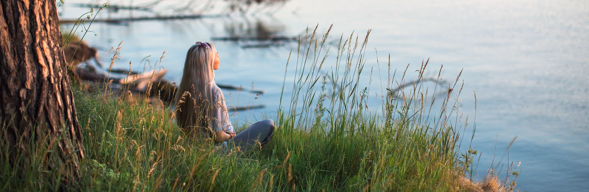Person with long hair sits on grass near a tree, looking out over calm water at sunset.